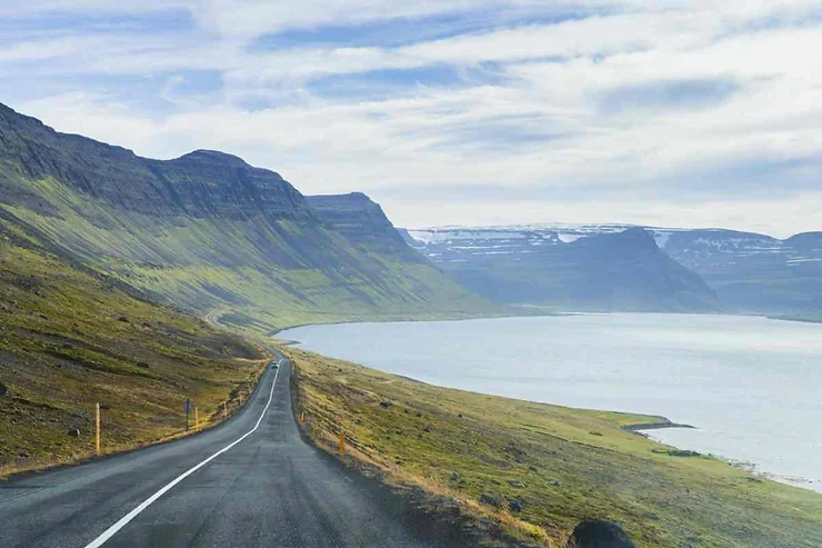 Scenic coastal road leading through the mountains of Seyðisfjörður, Iceland, with a view of the fjord and distant snow-capped peaks.