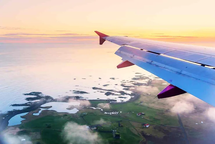 A stunning aerial view from an airplane window, showing the wingtip against a backdrop of a soft, pastel-colored sky during sunrise or sunset. Below, the coastline of Seydisfjordur, Iceland, is visible with its scattered lakes and lush green fields, partially covered by low-hanging clouds, merging with the tranquil ocean.