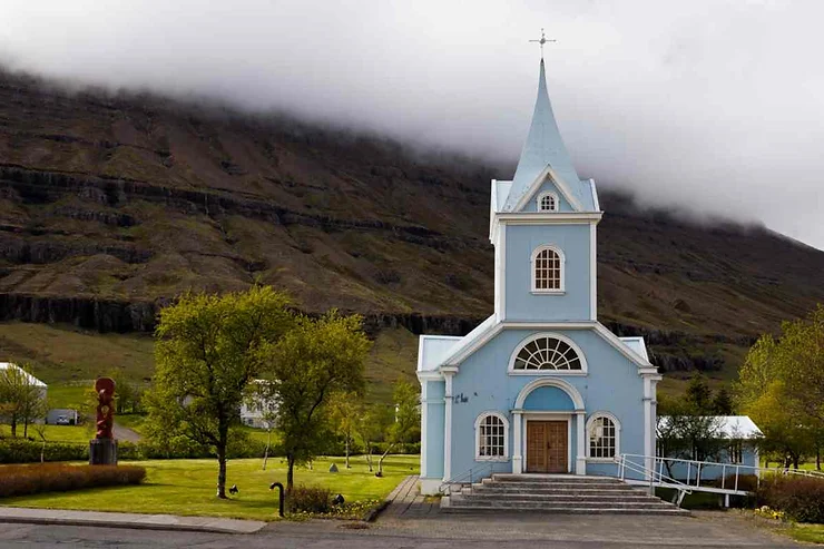 A serene view of the iconic blue church in Seydisfjordur, Iceland, set against a dramatic backdrop of mist-covered mountains. The quaint church, with its pointed steeple and arched windows, is surrounded by lush greenery, enhancing its tranquil and picturesque setting.