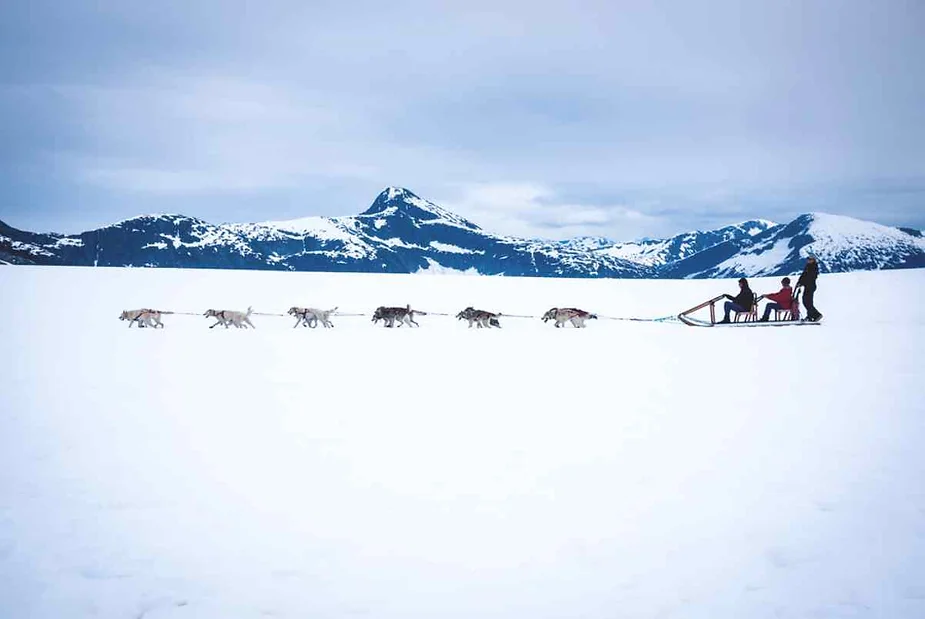 Dog sledding team pulling a sled with two passengers and a musher across a snowy landscape with mountains in the background, under a cloudy sky