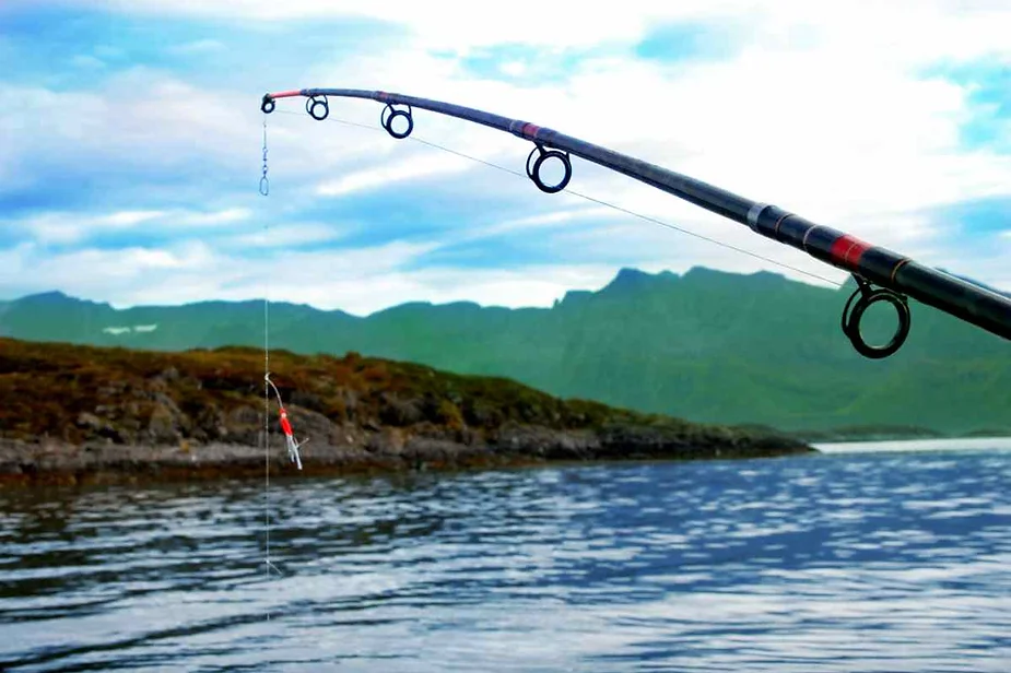 Fishing rod with a lure hanging over a calm lake, surrounded by green hills and mountains under a cloudy sky, capturing a serene fishing scene