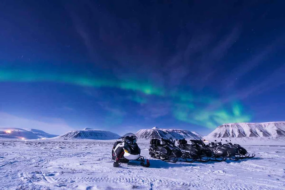 Group of snowmobiles parked on a snowy landscape in Iceland, with the Northern Lights illuminating the night sky and snow-covered mountains in the background