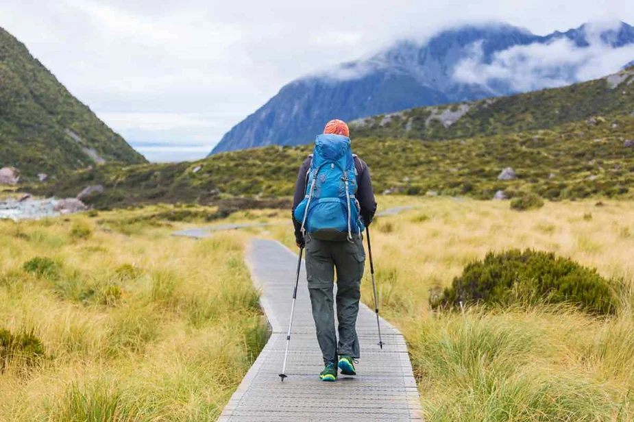 Hiker with a large blue backpack and trekking poles walking on a boardwalk trail through grassy terrain, surrounded by mountainous landscapes and misty clouds