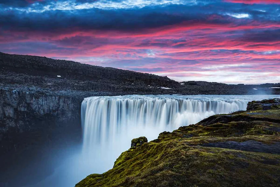 Majestic waterfall in Iceland with powerful water flow cascading over a cliff, surrounded by rocky terrain and vibrant, colorful skies during sunset