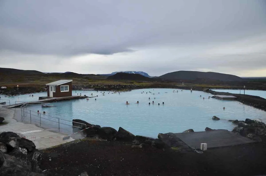 Natural geothermal lagoon in Iceland with milky blue water, surrounded by rocky terrain and mountains in the distance, with people enjoying the warm water