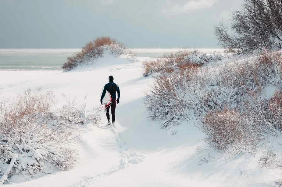 Person dressed in winter gear walking through snow-covered landscape with shrubs and dunes, holding a red sled, under a cloudy sky