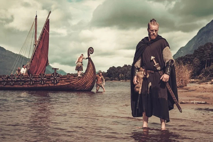 A Viking reenactor dressed in traditional garb stands in shallow water with a longship in the background and other reenactors aboard and onshore.