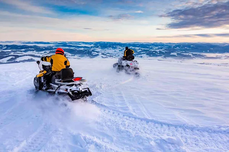 Two people riding snowmobiles across a snowy landscape with mountains in the background near Vik, Iceland.