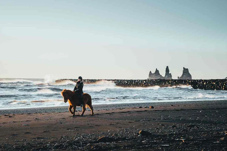 A rider on horseback traverses the black sand beach of Vík, Iceland, with the iconic Reynisdrangar sea stacks visible in the background.