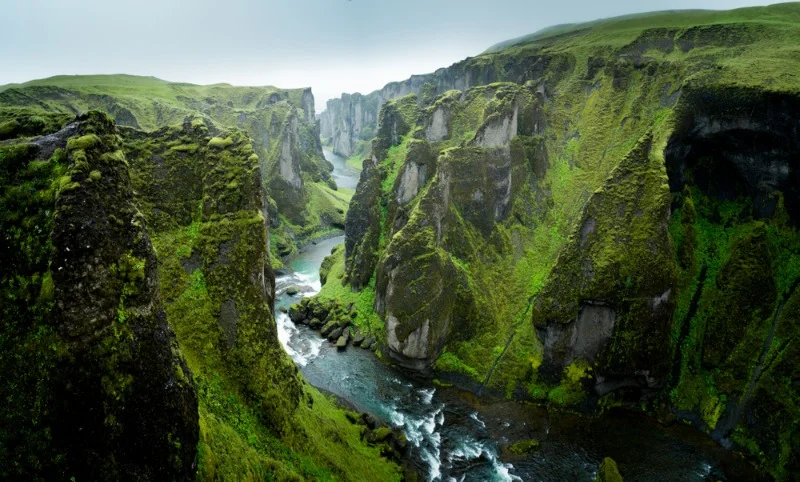 Breathtaking view of Fjaðrárgljúfur Canyon in Iceland, with lush green cliffs, a winding river, and a dramatic landscape shrouded in mist