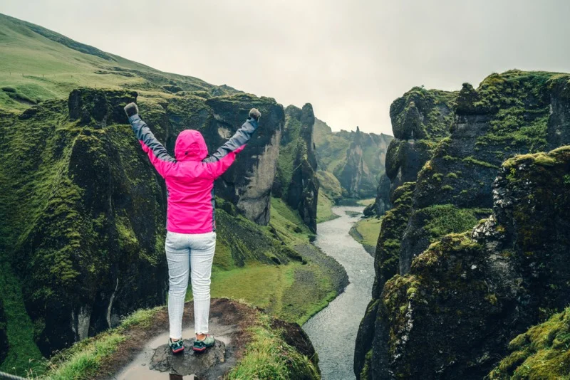 Traveler in a bright pink jacket celebrates atop a cliff at Fjaðrárgljúfur Canyon, Iceland, overlooking the lush green landscape and winding river below