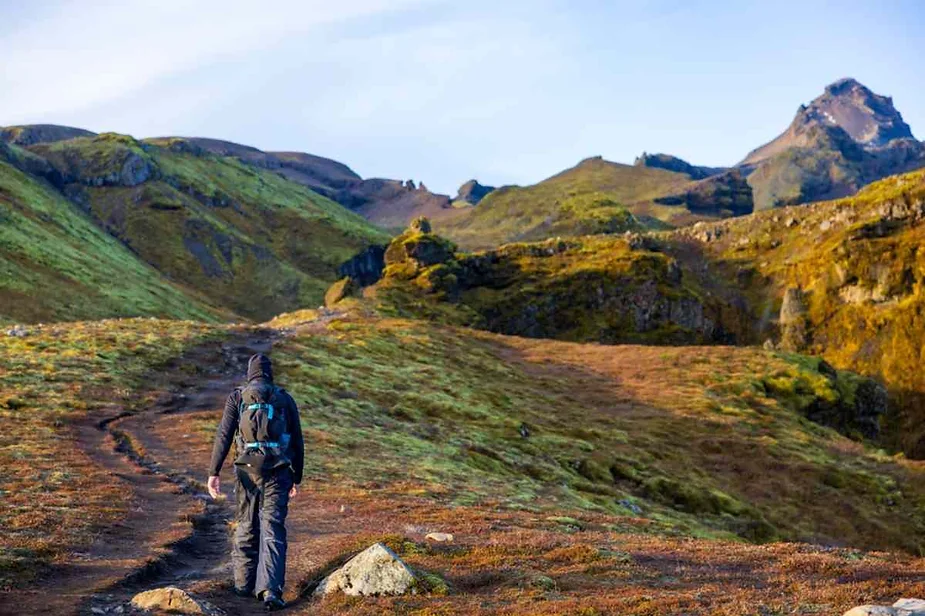 Solo hiker with a backpack walking along a rugged mountain trail surrounded by green hills and rocky terrain under a clear sky.