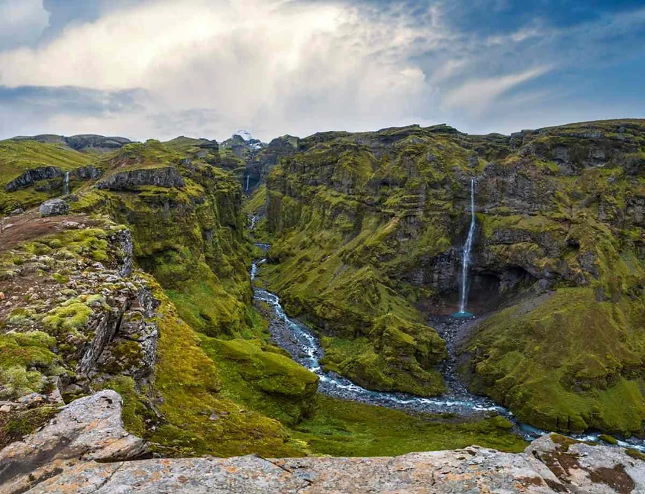 Expansive canyon landscape with vibrant green moss, a flowing river, and two cascading waterfalls under a partly cloudy sky.