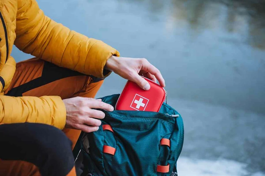 Hiker in a yellow jacket placing a red first aid kit with a white cross into a green backpack during an outdoor adventure.