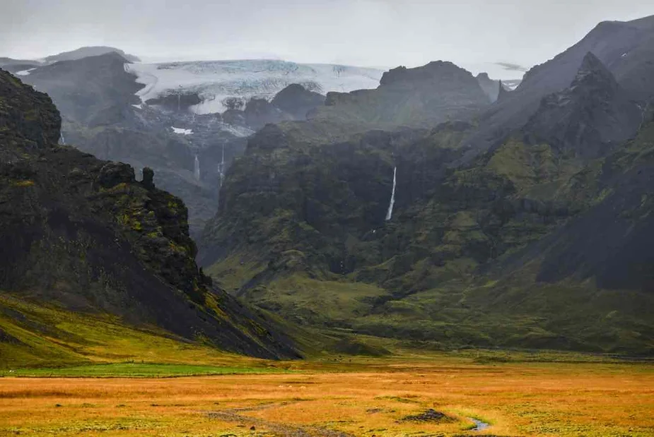 Vast, open field with golden grass leading to towering, rugged mountains and a distant glacier with waterfalls cascading down rocky cliffs.