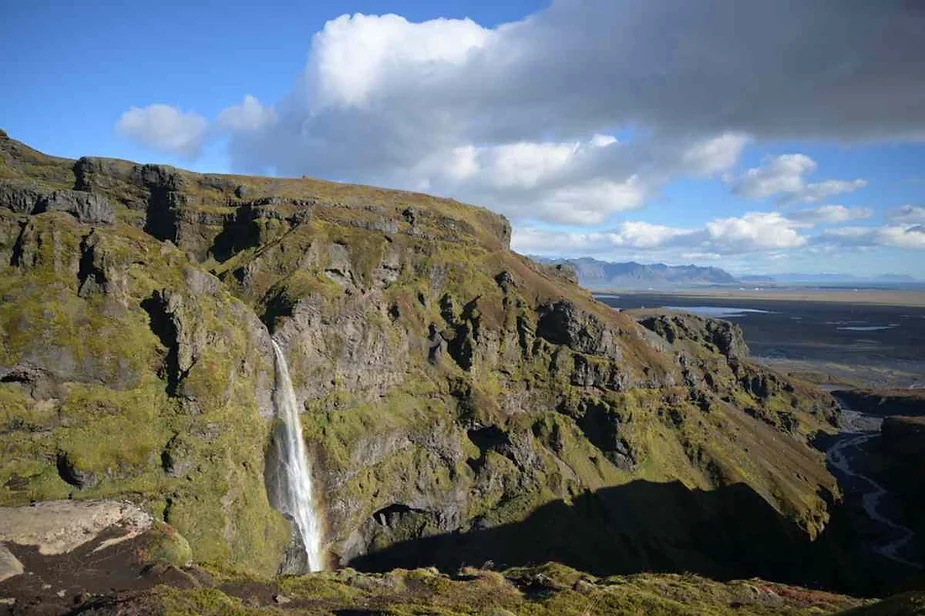 Scenic cliffside waterfall cascading down rugged rock formations with expansive plains and distant mountain range under a bright blue sky.