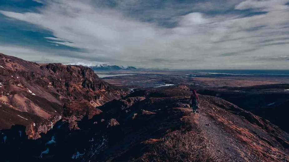 Solo hiker standing on a mountain ridge overlooking a vast valley and distant mountain range under a dramatic cloudy sky.