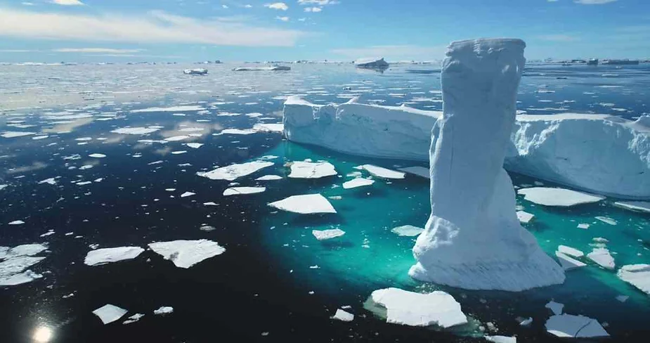 Massive icebergs floating in deep blue Arctic waters with patches of melting sea ice under a clear, sunny sky.