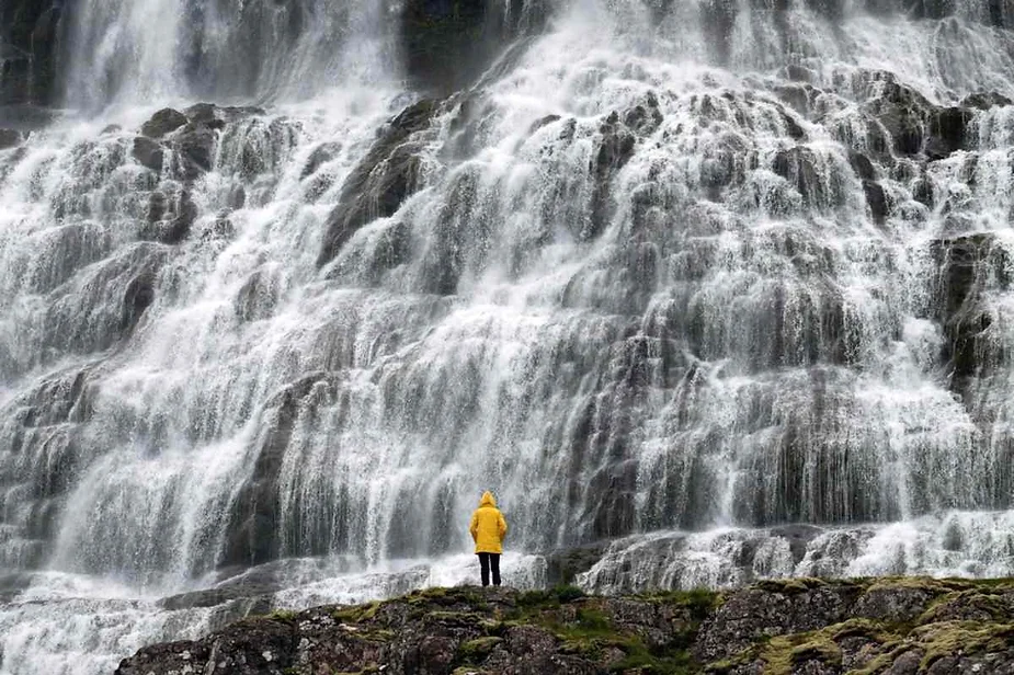 Person in a yellow raincoat standing at the base of a massive, cascading waterfall, surrounded by rocky terrain.