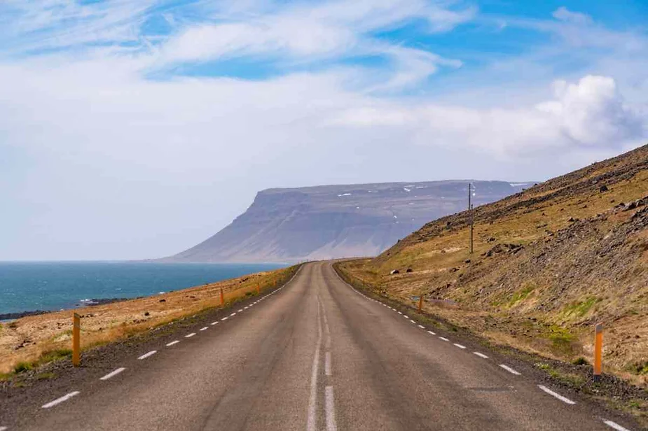 Long open road stretching towards a distant mountain along a coastal landscape under a bright blue sky.