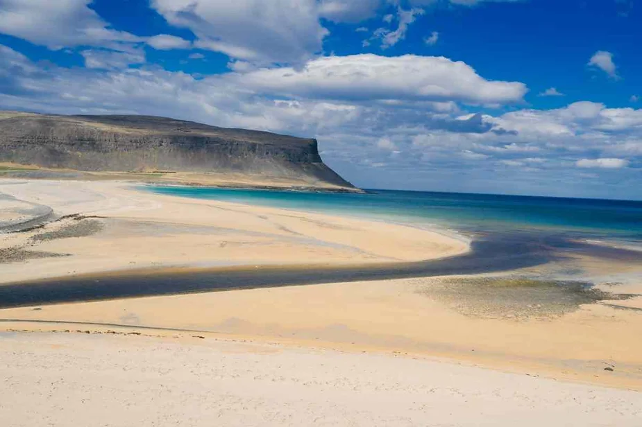 Pristine sandy beach with turquoise waters and a dramatic cliffside under a vibrant blue sky with scattered clouds.