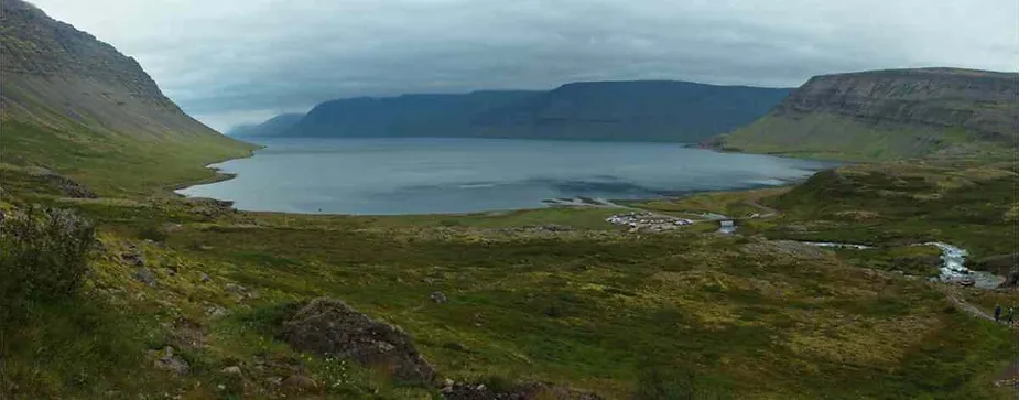 Wide panoramic view of a tranquil fjord surrounded by steep, green hills and distant mountains under a cloudy sky.