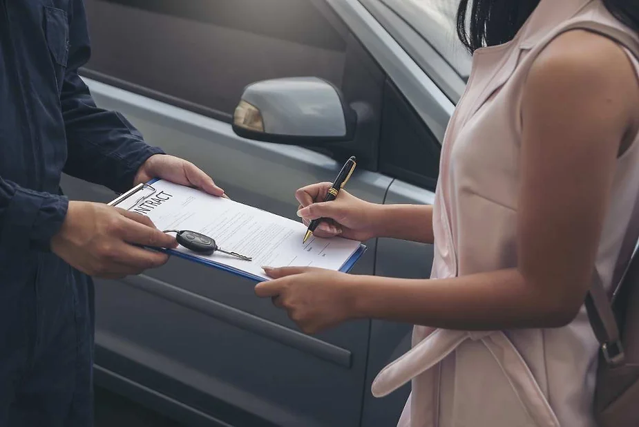 A woman signing a car insurance contract on a clipboard, held by a car dealership representative, with a car in the background