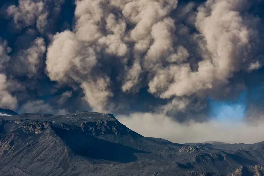 Massive volcanic ash cloud rising above a mountain during an eruption in Iceland, with dramatic and dark atmospheric conditions