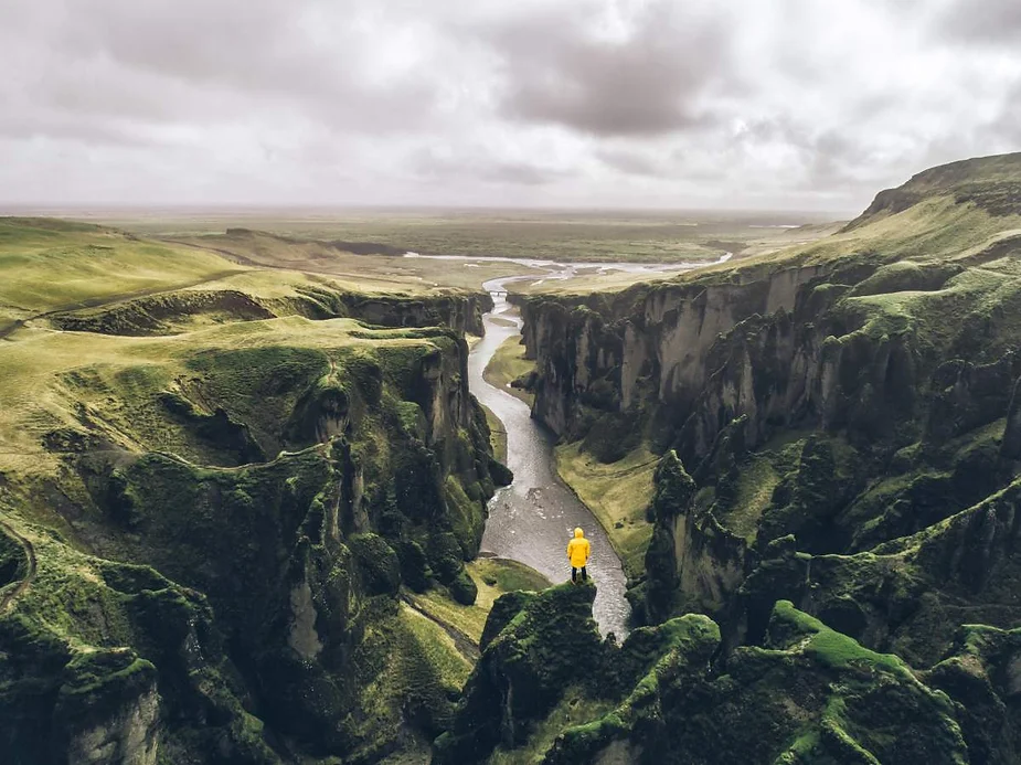 Aerial view of a person in a yellow jacket standing on a cliff edge overlooking the green and rugged Fjaðrárgljúfur Canyon with a winding river below