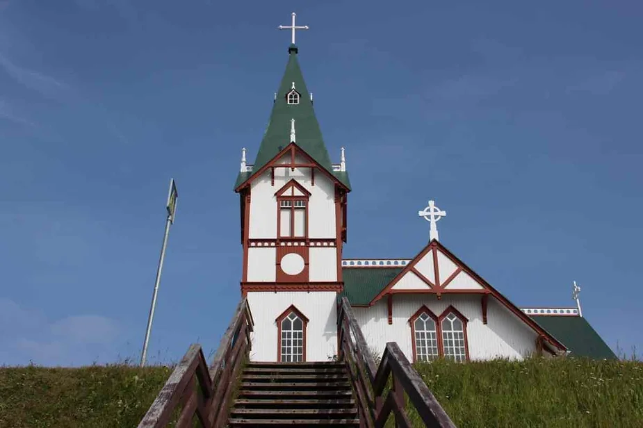 Historic wooden church with green and white details, standing on a hill under a clear sky in Húsavík, Iceland.