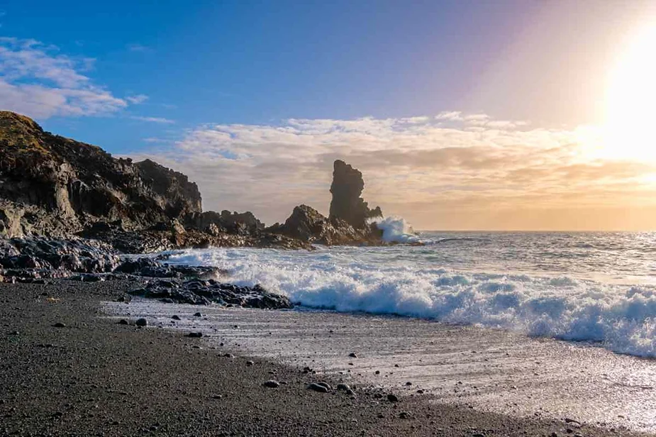 Sunset over a black sand beach in Iceland with crashing waves and a large sea stack rising from the rocky coastline.