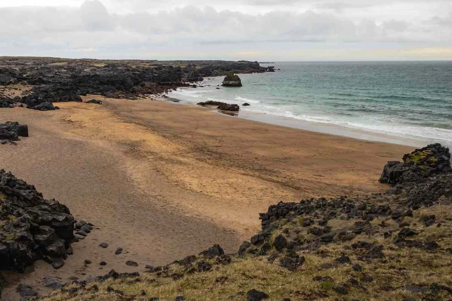 Secluded sandy beach surrounded by black volcanic rocks along the rugged coast of Iceland, with calm waves under a cloudy sky.