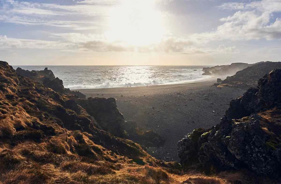 Sun setting over a secluded black sand beach in Iceland, framed by rugged cliffs and rocky coastline.