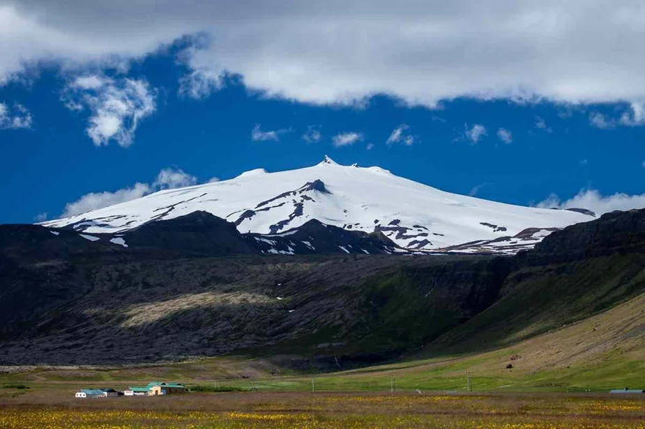 Snow-covered Snæfellsjökull volcano in Iceland, towering above green fields and small farmhouses under a partly cloudy sky.