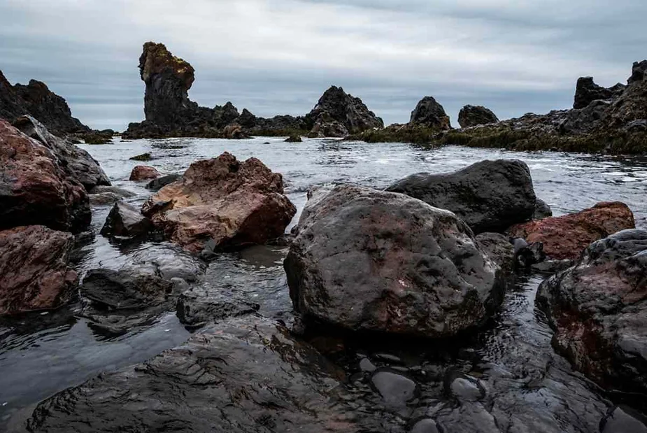 Rocky shore with large volcanic rocks and sea stacks rising from the water on Iceland’s rugged coastline under a cloudy sky.