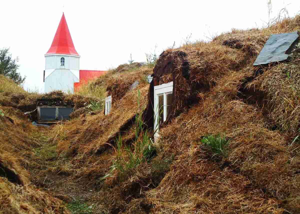 Traditional Icelandic turf houses with grass-covered roofs, accompanied by a white church with a red roof in the background