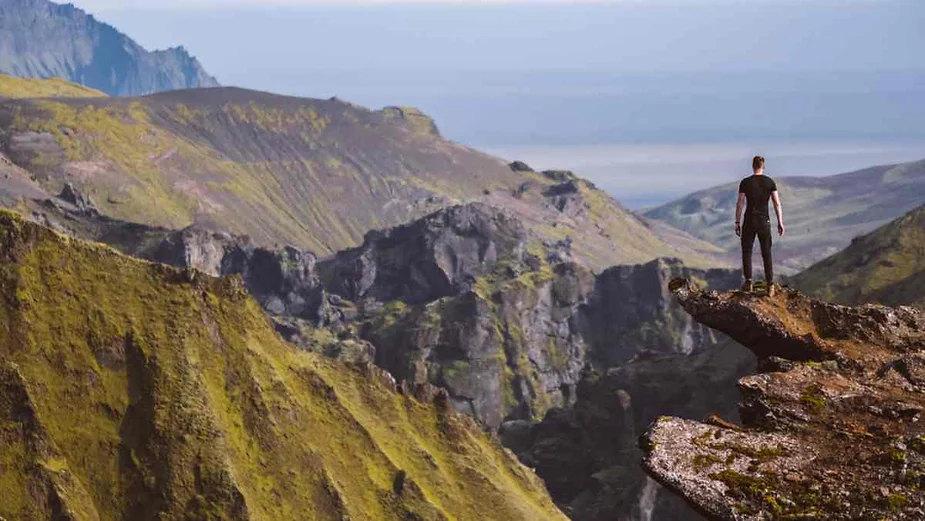 Man standing on a rocky cliff edge overlooking a vast Icelandic mountain range, surrounded by rugged green and gray landscapes under a clear sky.