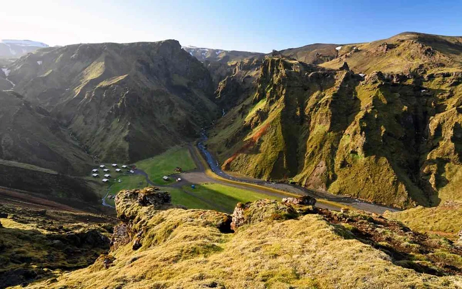 Aerial view of a scenic valley in Iceland with rugged cliffs, a winding road, and a small campsite nestled in the green landscape under a clear blue sky.