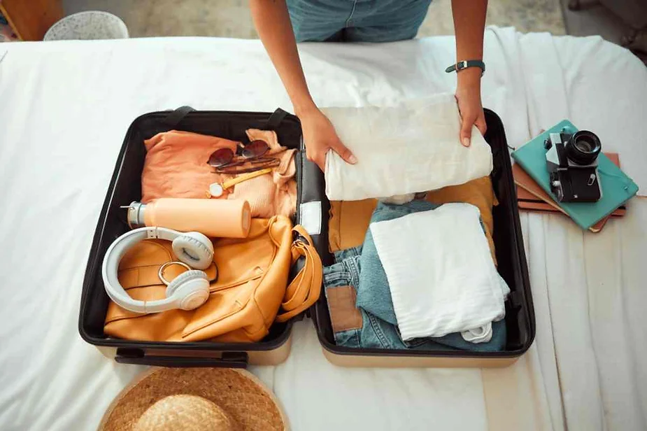 Person packing a suitcase with clothes, headphones, a water bottle, sunglasses, and a camera, preparing for a trip with travel essentials laid out.