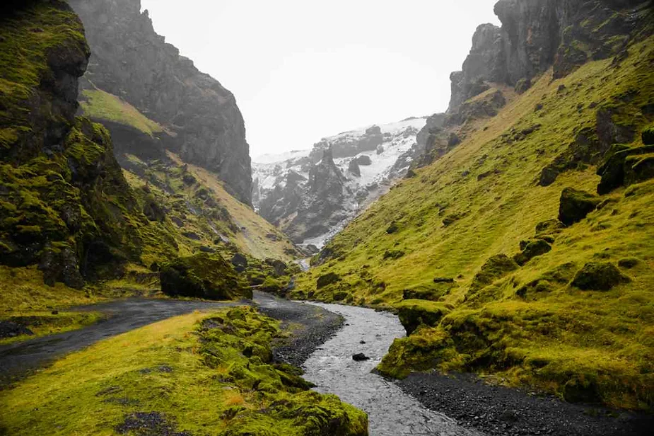 Narrow winding stream flowing through a moss-covered canyon in Iceland, with steep rocky cliffs and distant snow-capped peaks.