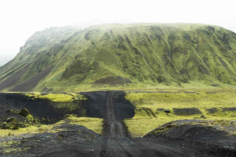 Dirt road cutting through a volcanic landscape in Iceland, leading towards a large green moss-covered hill shrouded in mist.