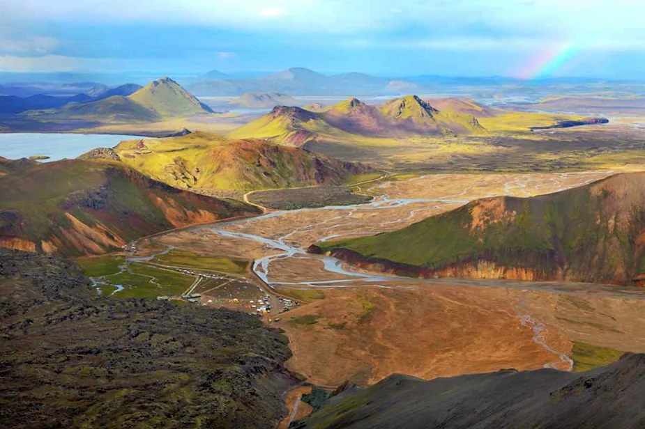 Vast aerial view of colorful Icelandic highlands with winding rivers, rugged volcanic hills, and a distant rainbow arching over the landscape.