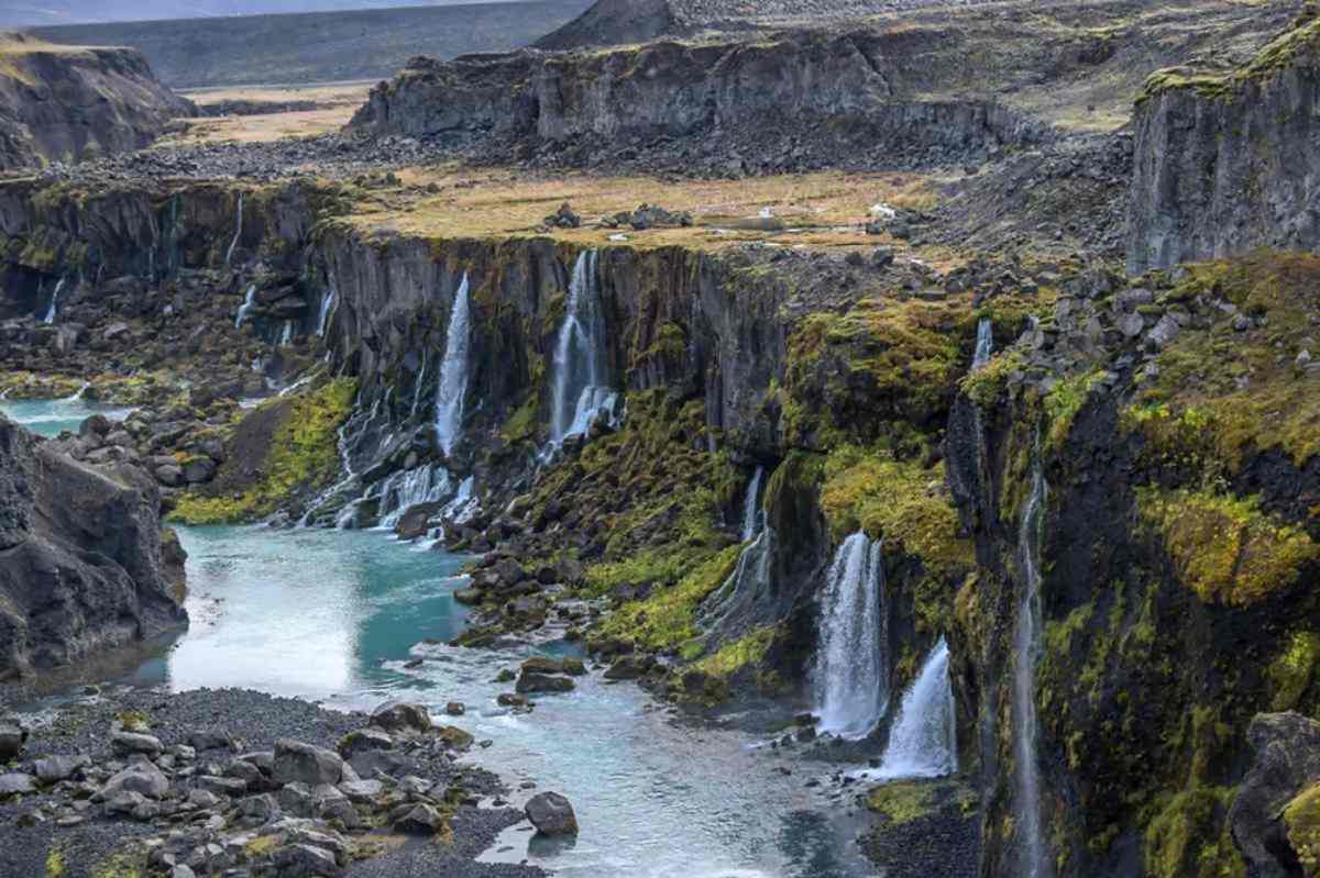 Series of cascading waterfalls flowing down moss-covered cliffs into a turquoise river in a remote Icelandic canyon surrounded by rugged terrain.