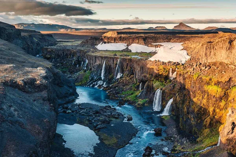 Scenic view of a rugged Icelandic canyon with multiple waterfalls cascading into a turquoise river, surrounded by snow patches and volcanic mountains.