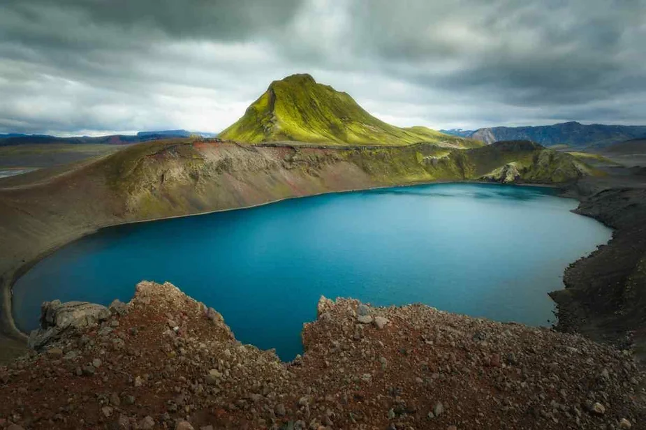 Vibrant turquoise lake nestled within a volcanic crater, surrounded by rugged terrain and a towering green mountain under a moody cloudy sky in Iceland.