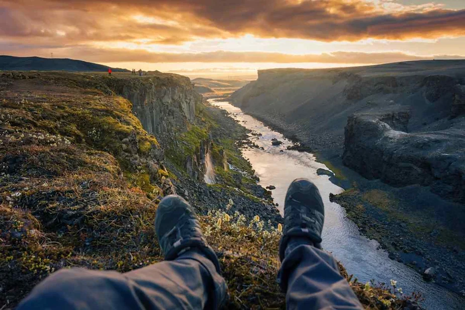 Person relaxing with feet stretched out, overlooking a scenic canyon with a winding river, steep cliffs, and a stunning sunset sky in Iceland.