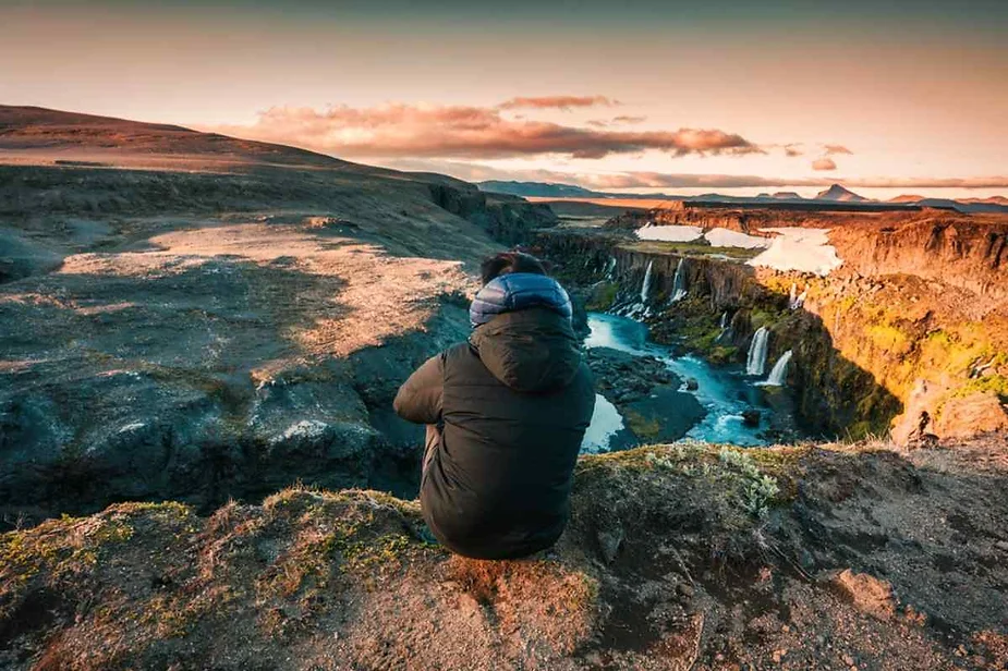 Traveler in a warm jacket sitting on a cliff edge, admiring the breathtaking view of cascading waterfalls and a turquoise river in Iceland at sunset.