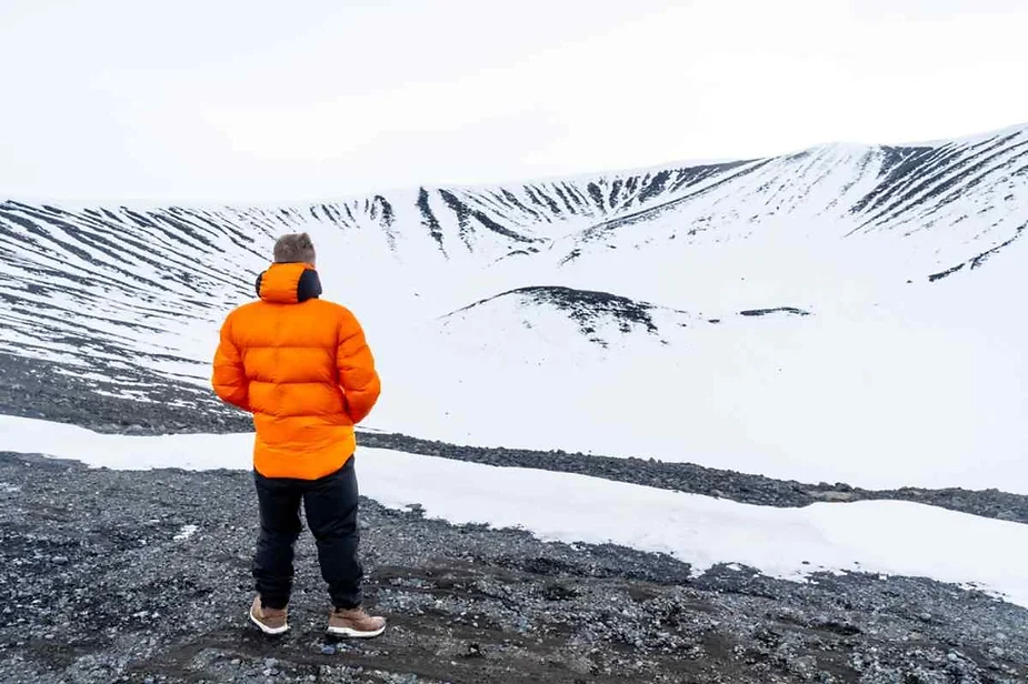 A man in an orange jacket stands at the snowy edge of Hverfjall Crater in Iceland, gazing into the vast, snow-covered volcanic crater under a cloudy sky.