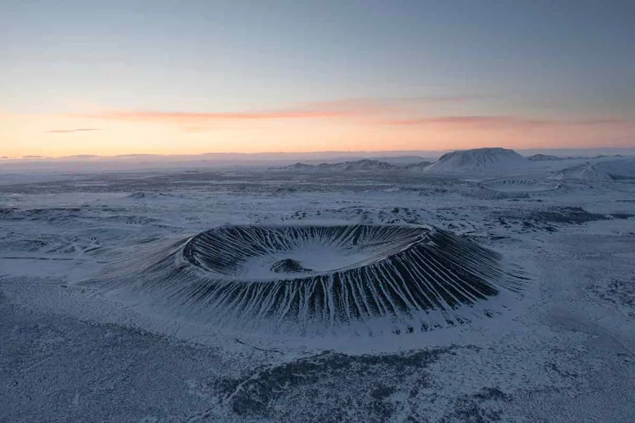 Aerial view of Hverfjall Crater in Iceland covered in snow at sunrise, with its dramatic volcanic slopes highlighted against the vast, frozen landscape and a soft pink sky.