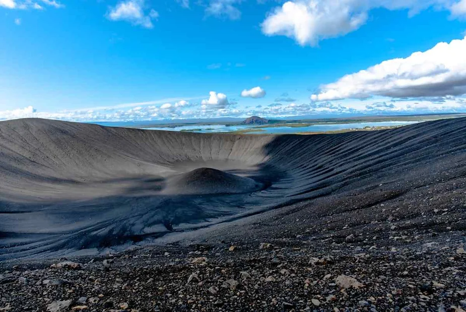 View from inside the Hverfjall Crater in Iceland, with dark volcanic slopes contrasting against a bright blue sky and scattered clouds, with distant lakes and mountains in the background.